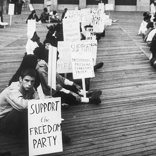 An integrated group of mostly young people sitting on the Atlantic City boardwalk. Many of them are holding signs showing support for the Mississippi Freedom Democratic Party (MFDP).